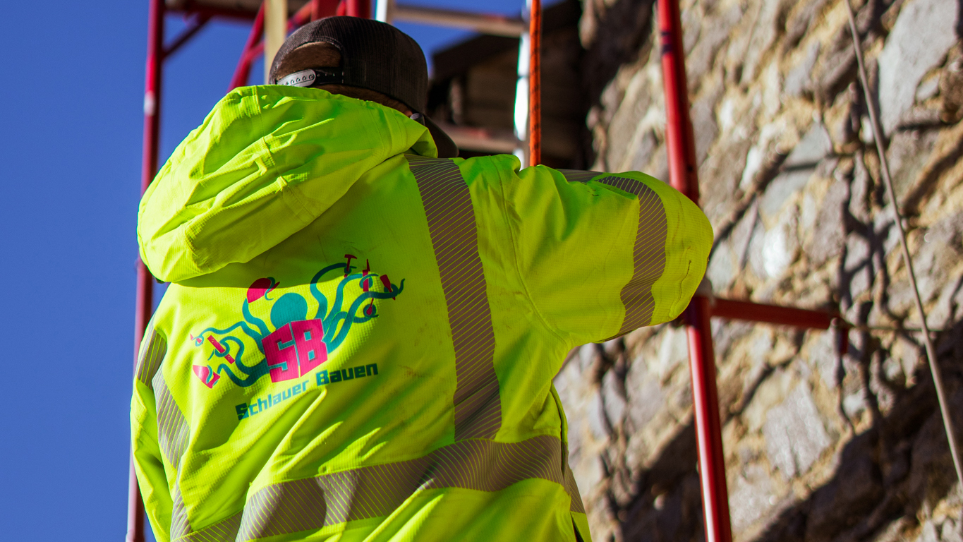 Ein Arbeiter von Schlauer Bauen während er in einem Gerüst der Burg Finstergrün. Er trägt eine leuchtend gelbe Warnjacke mit dem Schlauer Bauen Logo auf dem Rücken.